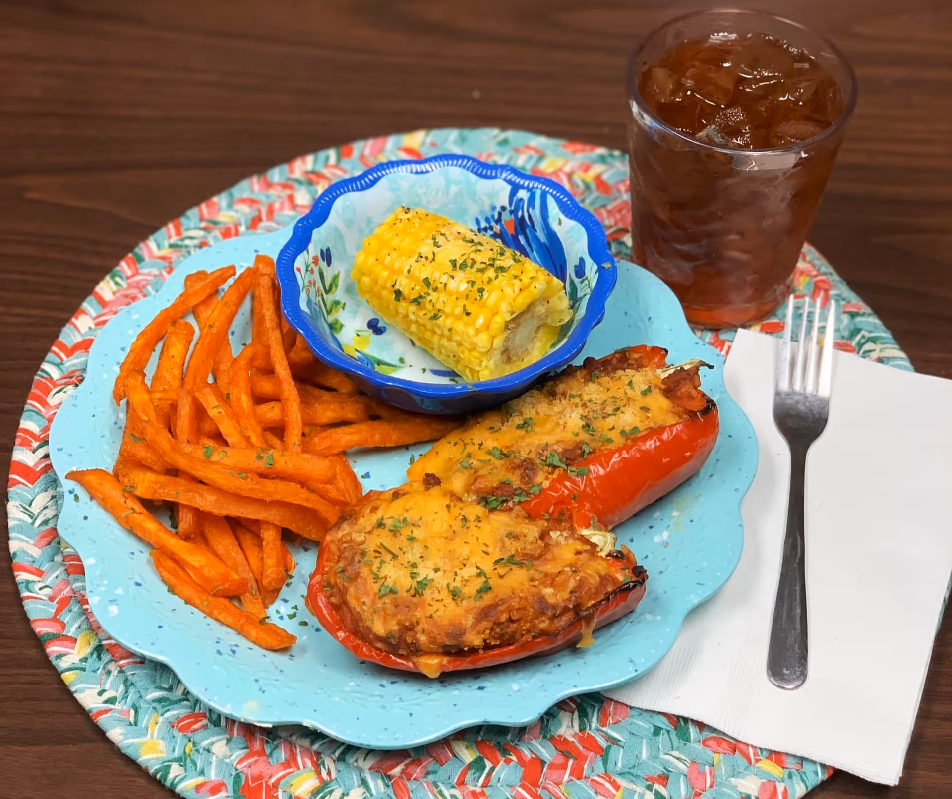 A meal on a blue plate with two stuffed red bell peppers topped with melted cheese and herbs, a serving of sweet potato fries, and a small bowl containing a piece of corn on the cob garnished with herbs. Next to the plate is a glass of iced tea with a fork resting on a white napkin. The plate is placed on a colorful woven placemat on a wooden table.