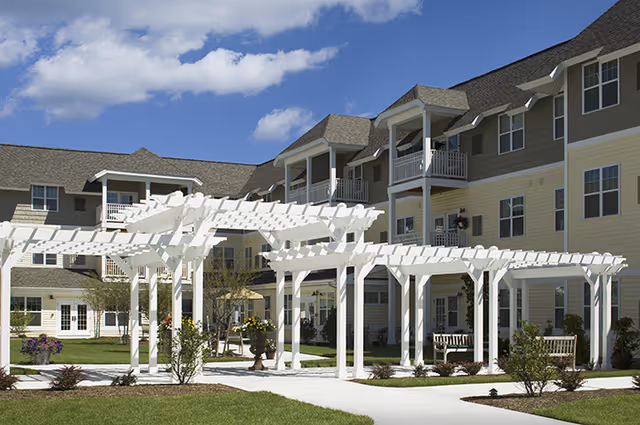 Outdoor courtyard area of a senior living facility with white wooden pergolas, benches, landscaped greenery, and a multi-story building with balconies in the background under a partly cloudy sky.