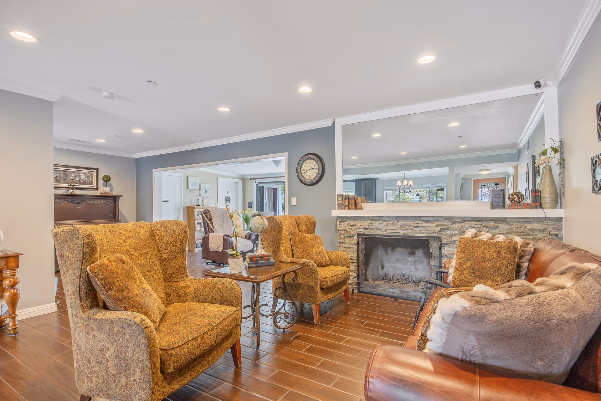 A cozy living room area with two patterned armchairs and a brown leather sofa with a fur throw. There is a wooden coffee table with a small plant and decorative items. The room features a stone fireplace with a large mirror above it, wooden flooring, and soft lighting from recessed ceiling lights. The walls are painted in neutral tones with some framed artwork and a clock.