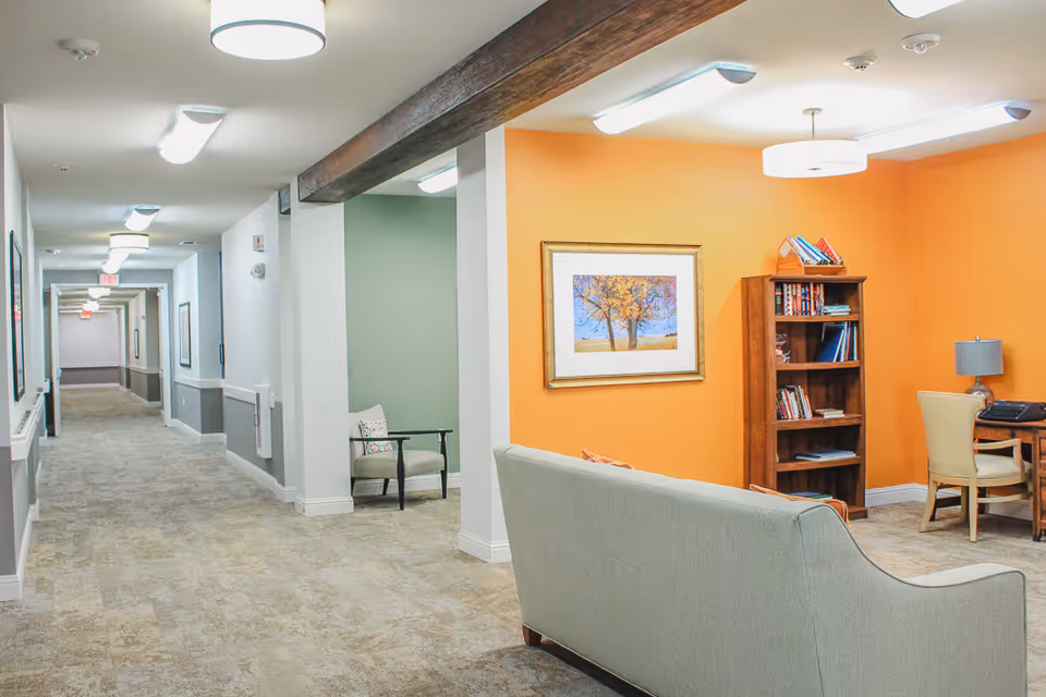 Interior view of a senior living facility hallway leading to a seating area with a light gray couch, a wooden bookshelf filled with books, a desk with a chair and a lamp, and an orange accent wall with a framed picture of a tree.