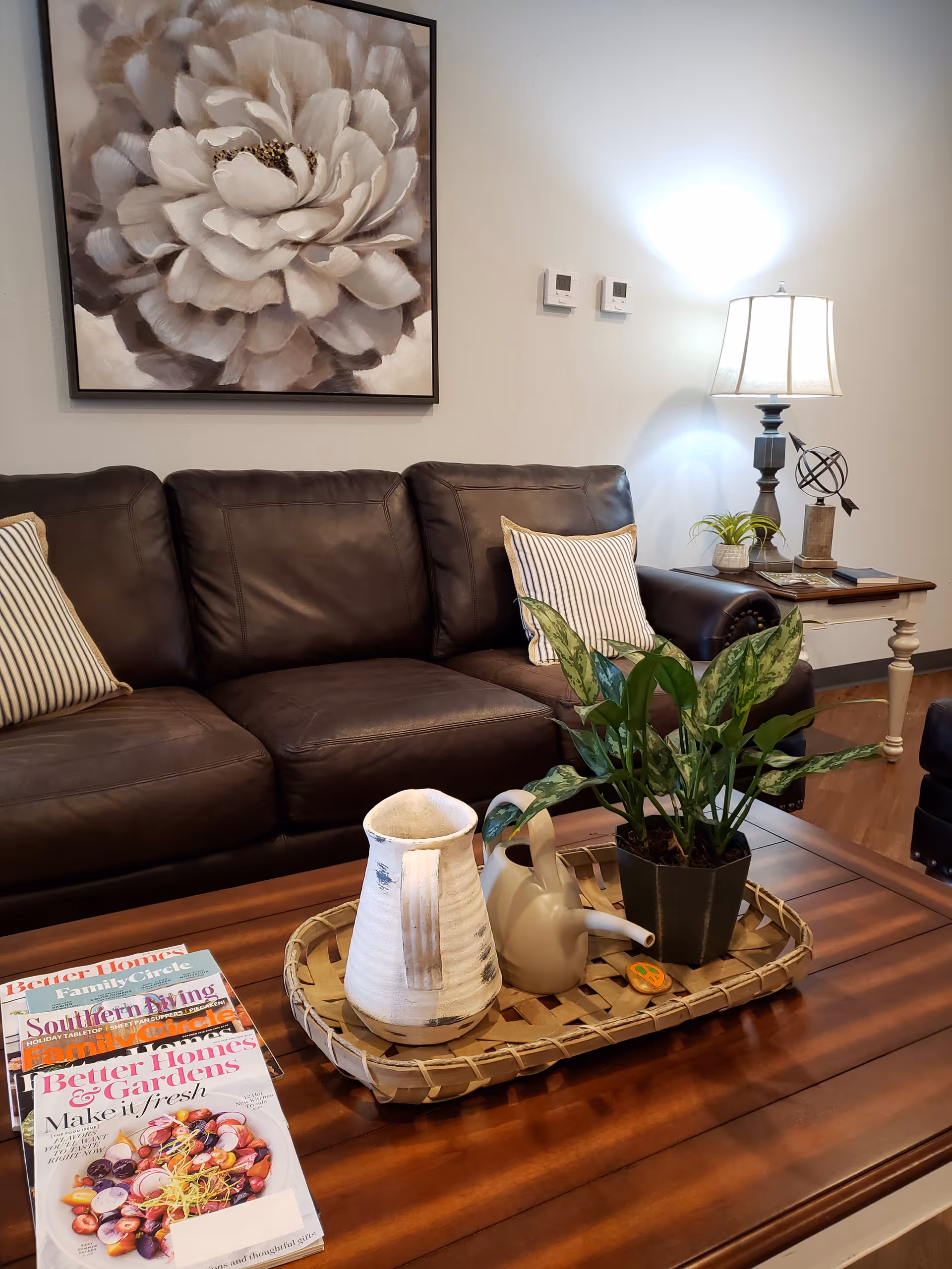A cozy living room area featuring a dark brown leather sofa with two striped pillows. Above the sofa hangs a large painting of a white flower. In front of the sofa is a wooden coffee table with a woven tray holding a white ceramic pitcher, a small watering can, and a potted green plant. To the right, there is a side table with a lamp, a small decorative globe, and a plant. Several magazines are stacked on the coffee table.