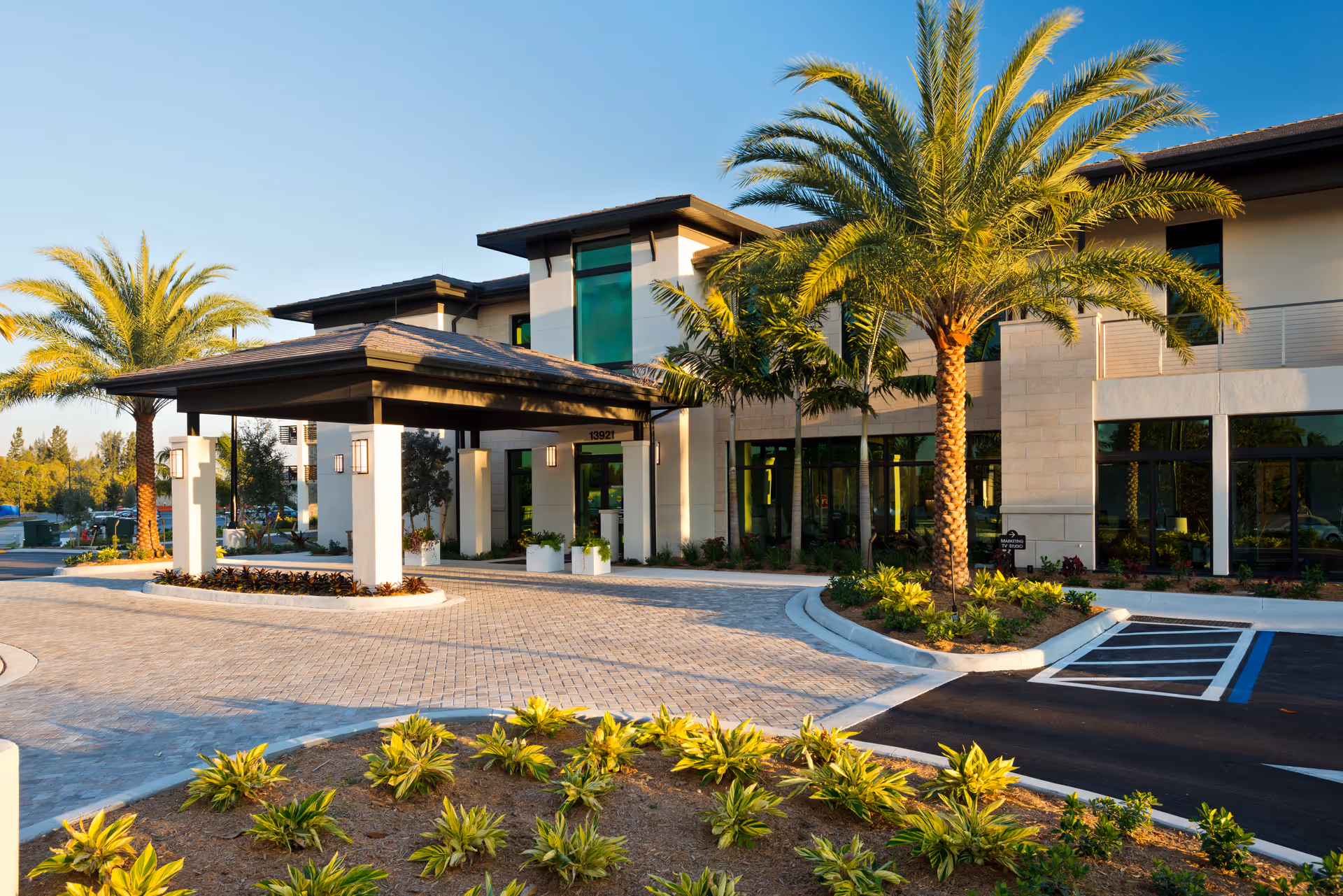 Exterior view of the Shell Point Welcome Center building with a covered entrance, palm trees, landscaped plants, and a paved driveway under a clear blue sky.