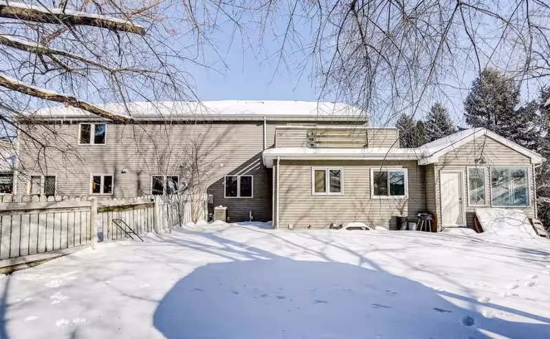 Exterior view of a two-story residential building with beige siding, surrounded by snow-covered ground and leafless trees under a clear blue sky.