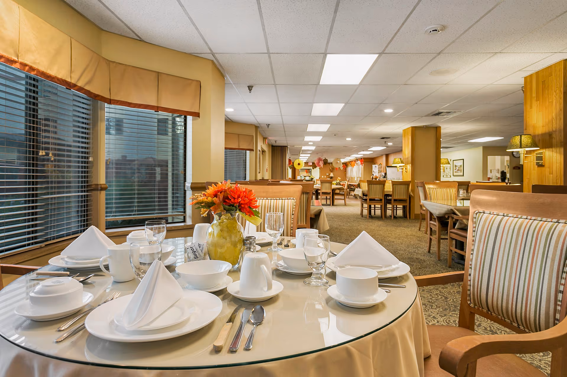 A dining room in a senior living facility with a round glass-top table set for a meal. The table has white plates, bowls, cups, silverware, and folded white napkins. A vase with orange and red flowers is in the center. The room has multiple tables and chairs, beige walls, large windows with blinds, and ceiling lights.