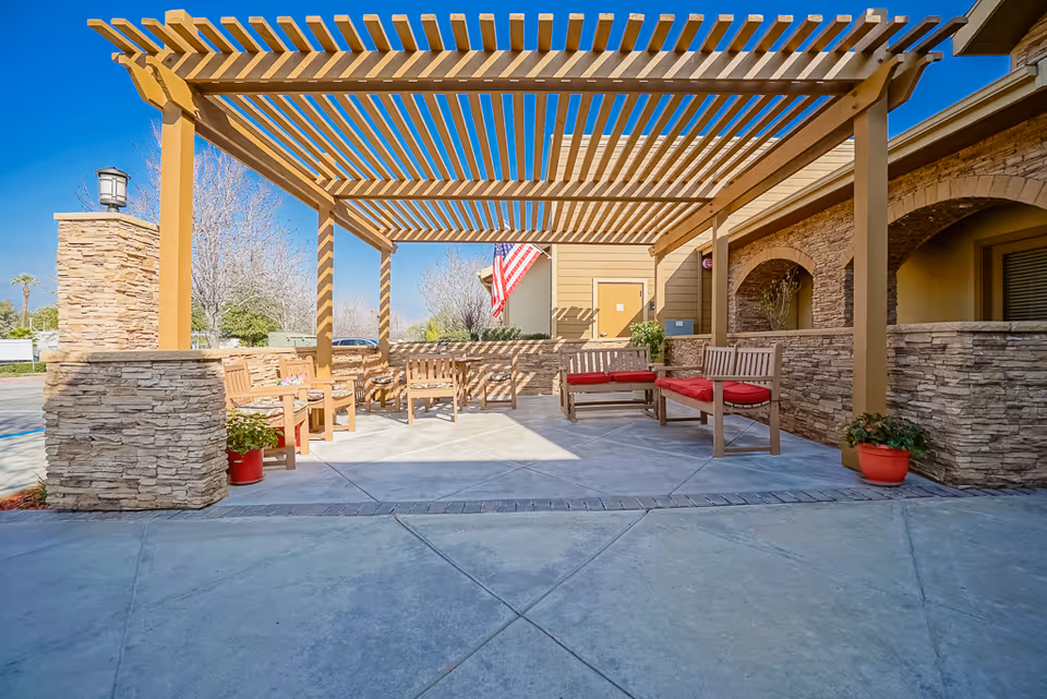 Outdoor seating area at Summerfield of Redlands with wooden benches featuring red cushions under a wooden pergola. Stone walls surround the space, and an American flag is visible in the background against a clear blue sky.