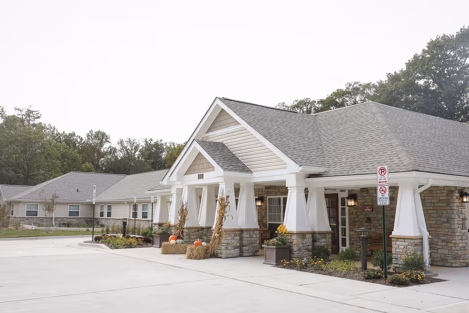Front entrance of a single-story senior living building with a gabled portico, stone pillars, pumpkins and landscaped beds.