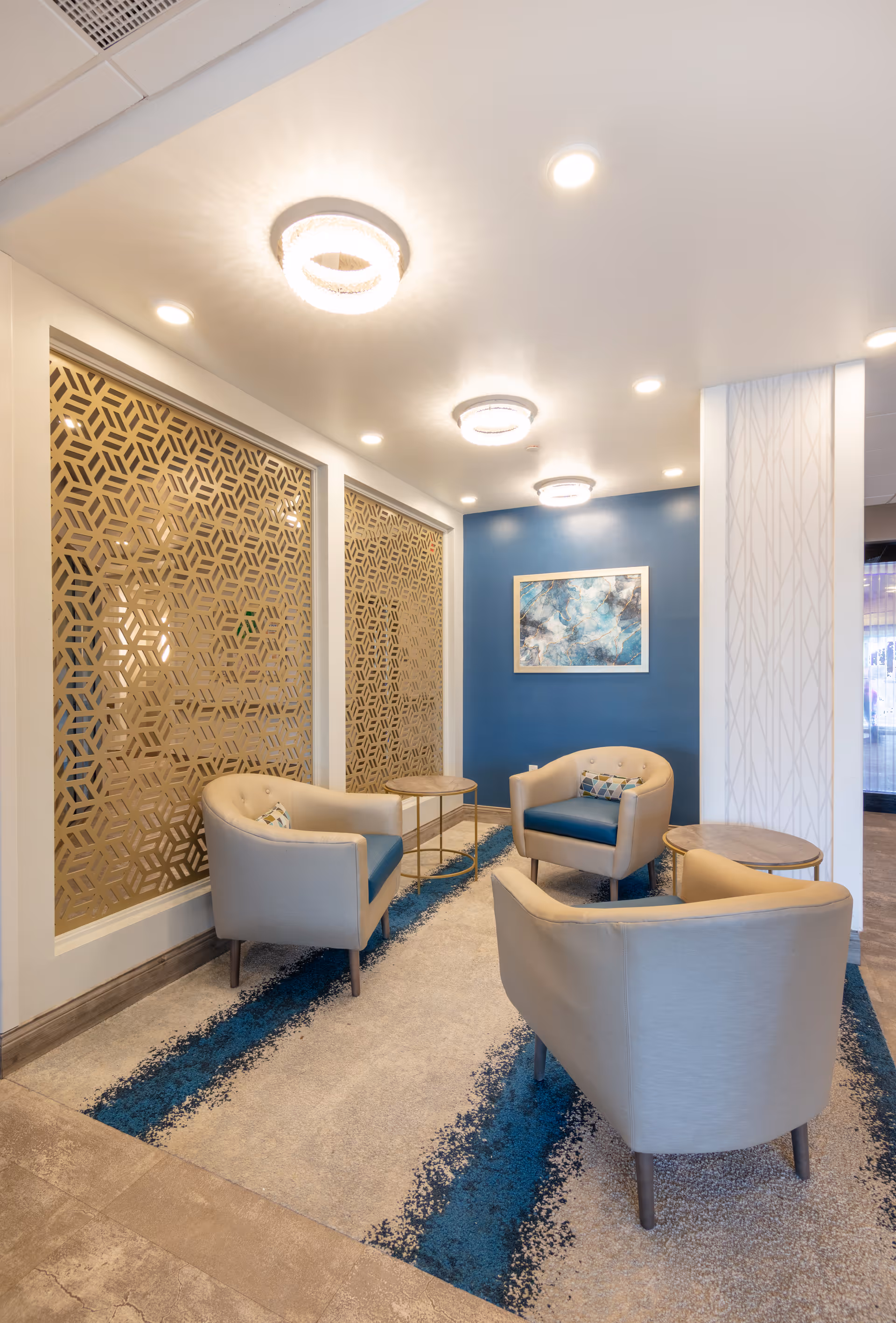 A modern sitting area with three beige armchairs featuring blue cushions arranged around two small round tables. The space has a blue accent wall with an abstract painting, decorative wooden lattice panels on one side, and a patterned carpet with blue and gray tones. The ceiling has recessed lighting and circular light fixtures.