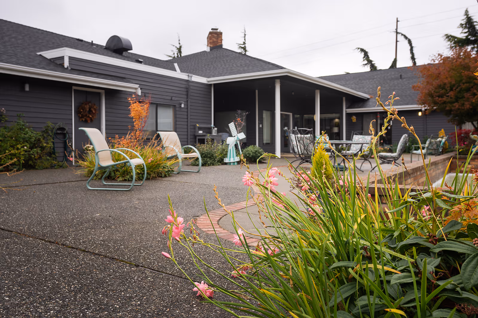 Outdoor patio area at Village Concepts of Oak Harbor - Harbor Tower Village featuring garden plants with pink flowers in the foreground, several chairs and tables arranged on a paved surface, and a dark gray building with a covered porch in the background.