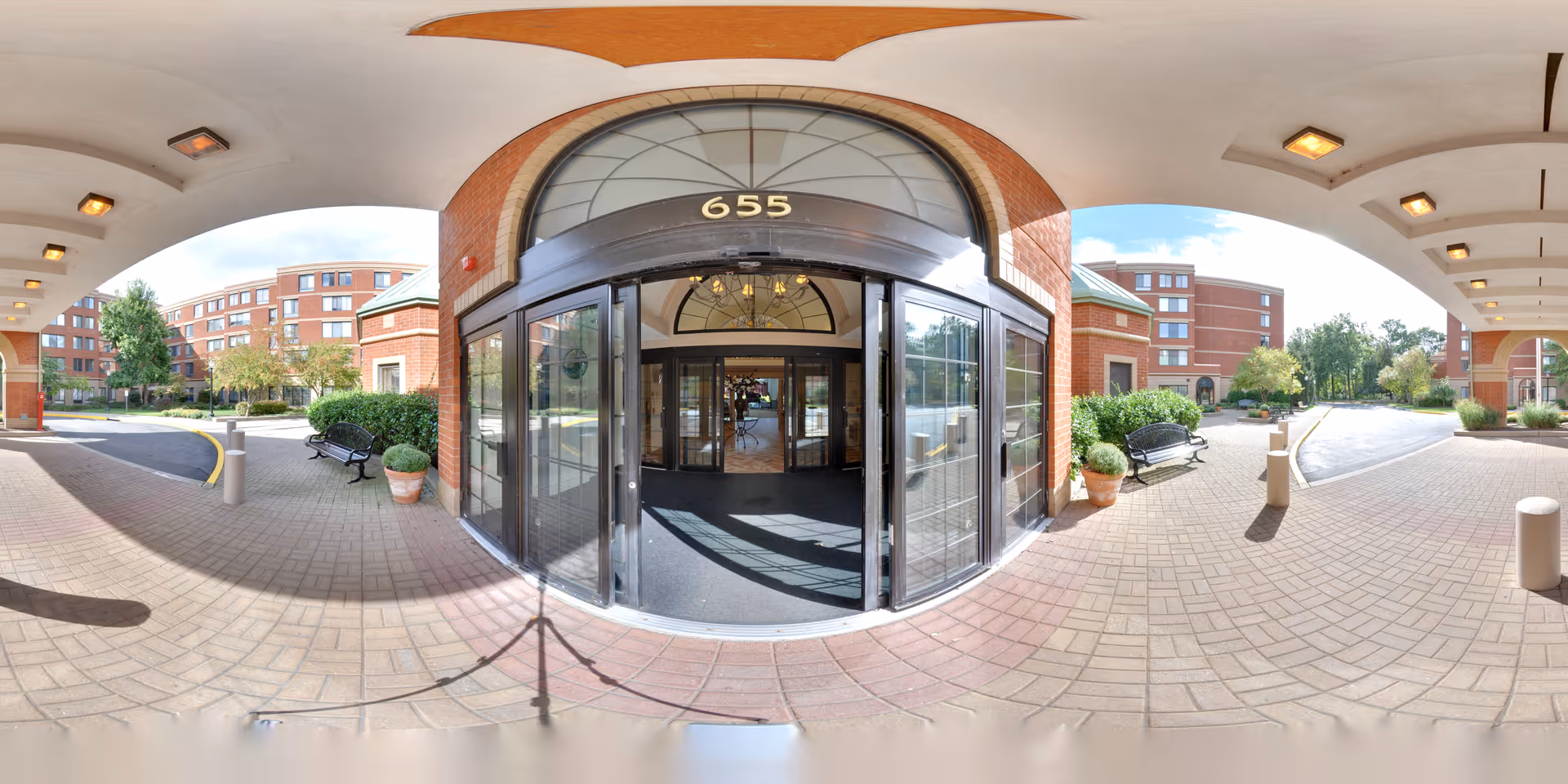 Entrance area of a senior living facility with a covered driveway and automatic glass doors marked with the number 655. The building is made of red brick with large windows. There are benches and potted plants on either side of the entrance, and a paved driveway curves around the front. Trees and greenery are visible in the background under a partly cloudy sky.