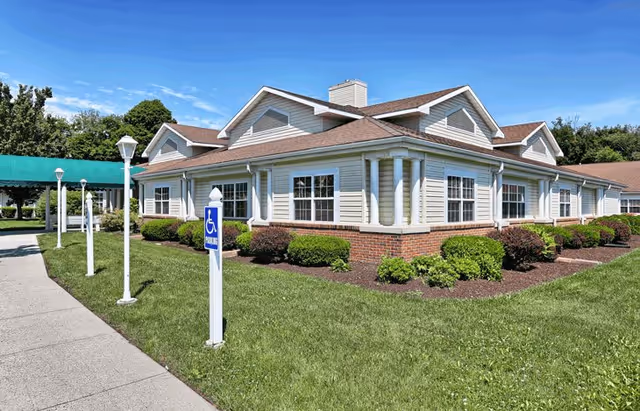 Exterior view of a single-story senior living facility building with beige siding and brick accents, surrounded by green grass and shrubs under a clear blue sky. A sidewalk with lamp posts and a handicap parking sign is visible in the foreground.