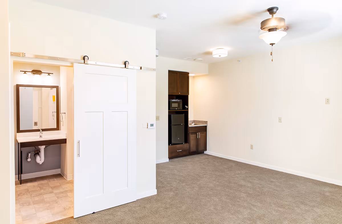 Carpeted studio living area with a kitchenette, ceiling fan, and a sliding barn door revealing a bathroom sink and mirror.