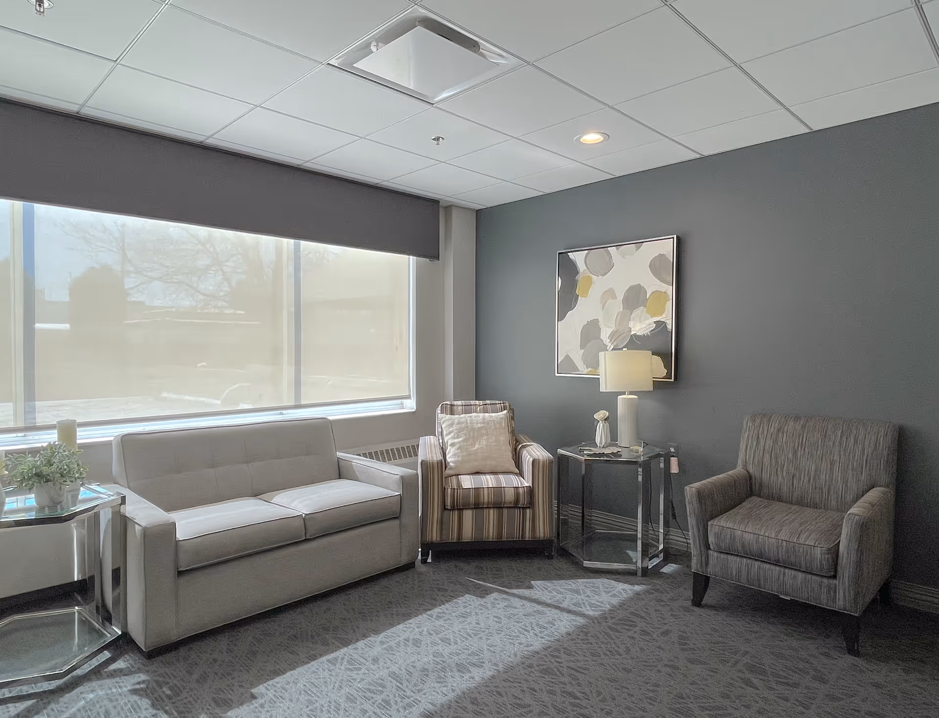 Sunlit seating area with a sofa and two armchairs arranged around glass side tables against a gray wall with artwork.