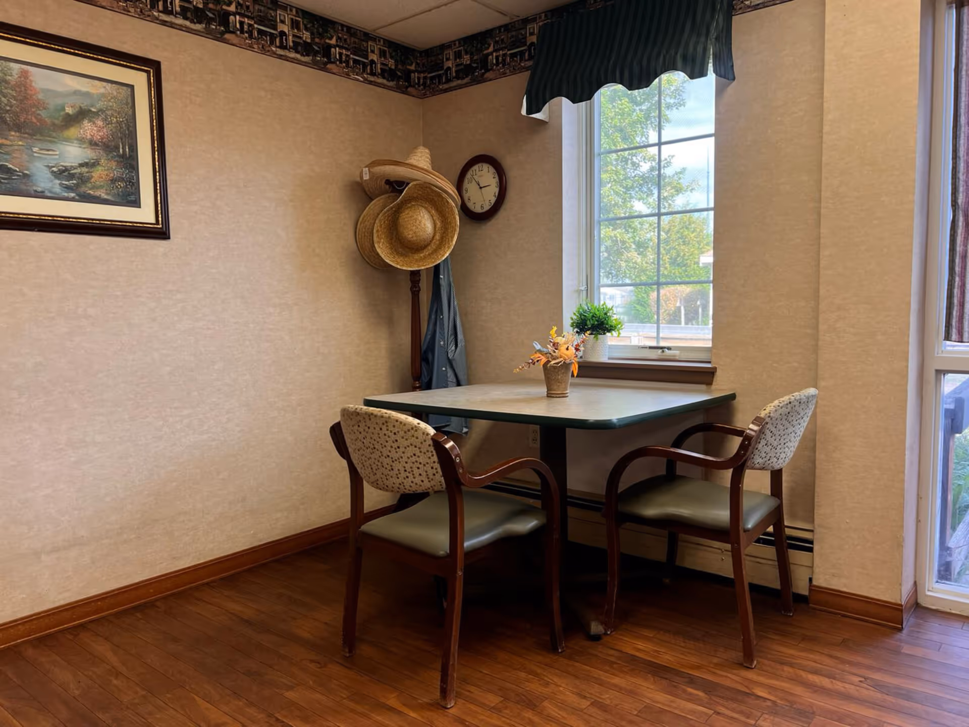 A small dining area with a square table and two chairs with patterned upholstery. On the table is a small flower arrangement. Behind the table is a window with a green valance, letting in natural light and showing trees outside. A coat rack with two straw hats and a hanging jacket is in the corner. A framed painting and a clock are mounted on the beige walls. The floor is wooden.