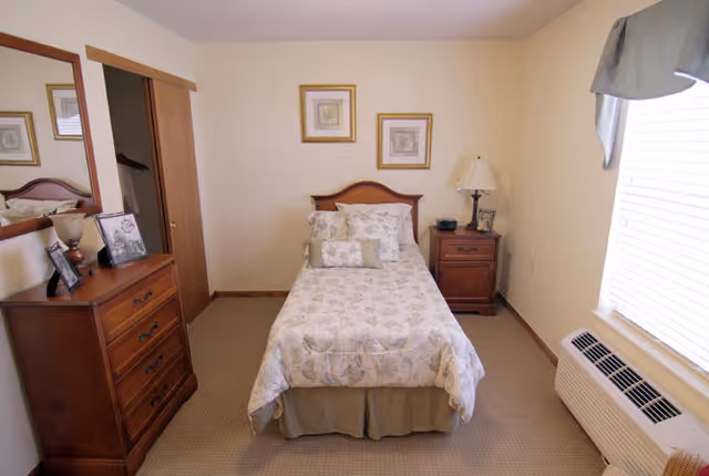 A small bedroom with a single bed centered against the back wall, covered with a floral patterned bedspread and multiple pillows. On the right side of the bed is a wooden nightstand with a lamp and a small clock. On the left side of the room is a wooden dresser with framed photos and a large mirror above it. There is a window with blinds and a valance on the right wall, and a closet with sliding wooden doors on the left wall.