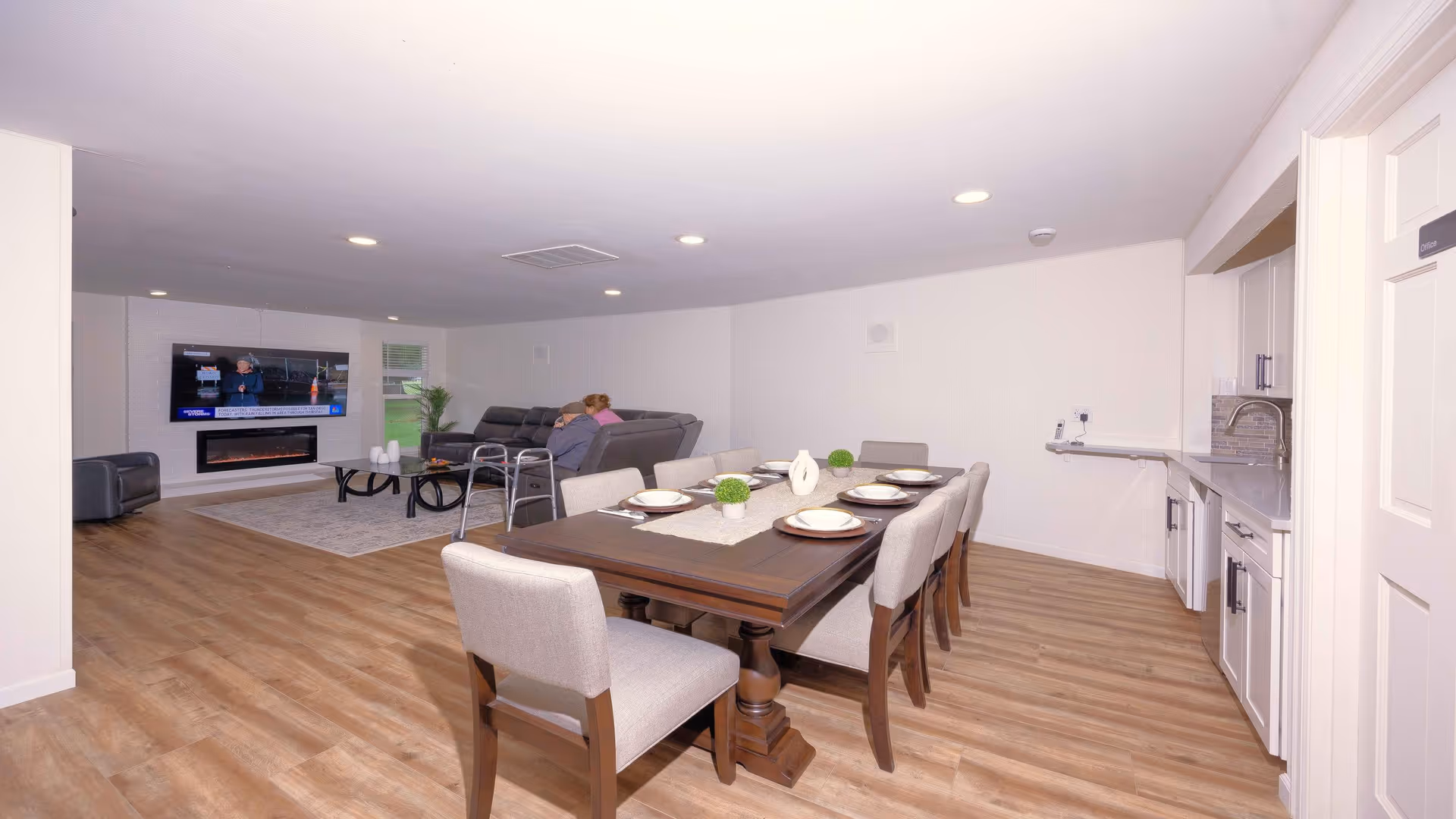 Open-plan dining area with a large wooden table set for six and a connected living room with a sofa and wall-mounted TV.