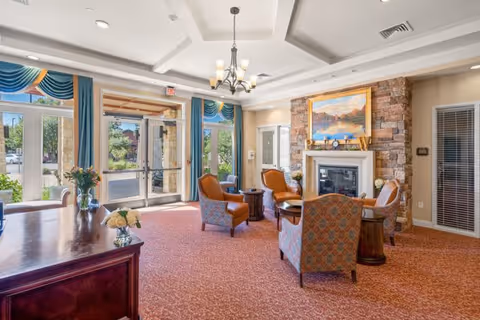 A bright and inviting common area with large windows and glass doors letting in natural light. The room features a stone fireplace with a painting above it, surrounded by four upholstered armchairs arranged around small wooden tables. The ceiling has a chandelier and decorative beams, and the floor is covered with a patterned red carpet. Blue and gold curtains frame the windows and doors, and a wooden table with flower vases is visible in the foreground.