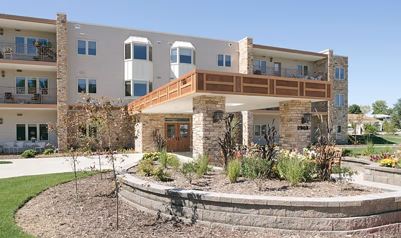 Exterior view of a three-story senior living facility with stone and beige siding, featuring balconies, a covered entrance supported by stone pillars, and landscaped garden beds with various plants and flowers in front.
