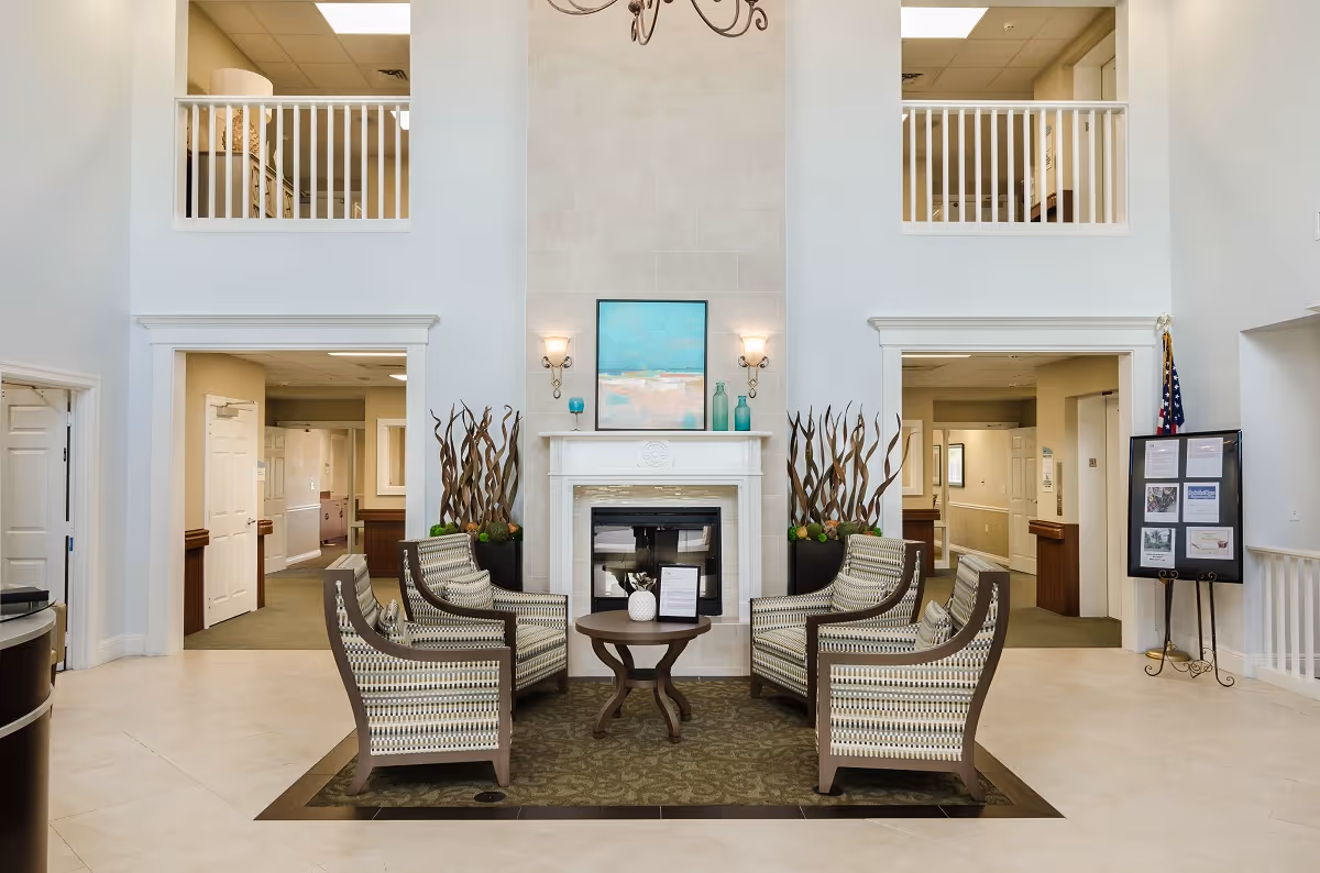 A bright and spacious common area in a senior living facility featuring a central fireplace with decorative vases and a painting above it. Four patterned armchairs are arranged around a small round table on a rug. The room has high ceilings with two open balcony-style railings above doorways on either side of the fireplace. There is a chandelier hanging from the ceiling and an American flag near a bulletin board on the right side.
