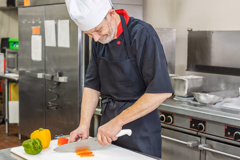 An elderly male chef wearing a white chef hat, black apron, and black shirt with red collar is slicing a red bell pepper on a white cutting board in a commercial kitchen. There are yellow and green bell peppers on the cutting board, and stainless steel kitchen equipment is visible in the background.
