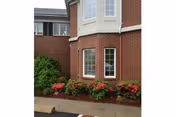 Exterior view of a red brick building with a bay window and flower beds containing red and pink flowers along the base. There is a sidewalk and part of a parking area visible in front of the building.