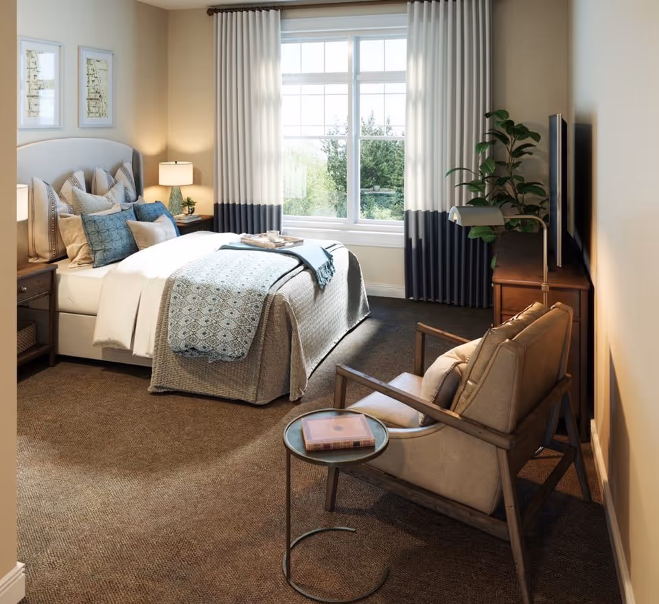 A cozy bedroom with a neatly made bed featuring multiple pillows and a patterned throw blanket. There is a wooden nightstand with a lamp and a small plant beside the bed. A large window with white and navy curtains lets in natural light. In the foreground, there is a cushioned armchair with a small round side table holding a book. A wooden dresser with a TV and a floor lamp is against the wall, and a tall green plant adds a touch of nature to the room.