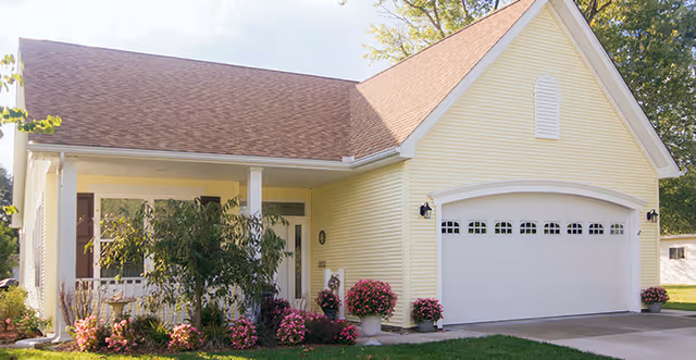 Exterior view of a single-story yellow house with a brown shingled roof, a white garage door, a small front porch with white railings, and a garden with pink flowers and green shrubs.