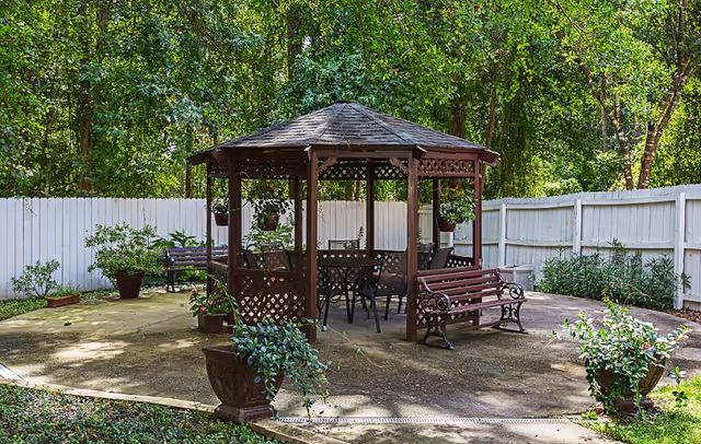 Outdoor seating area with a wooden gazebo surrounded by a white fence and greenery. The gazebo has a table and chairs inside, with benches and potted plants around the concrete patio.