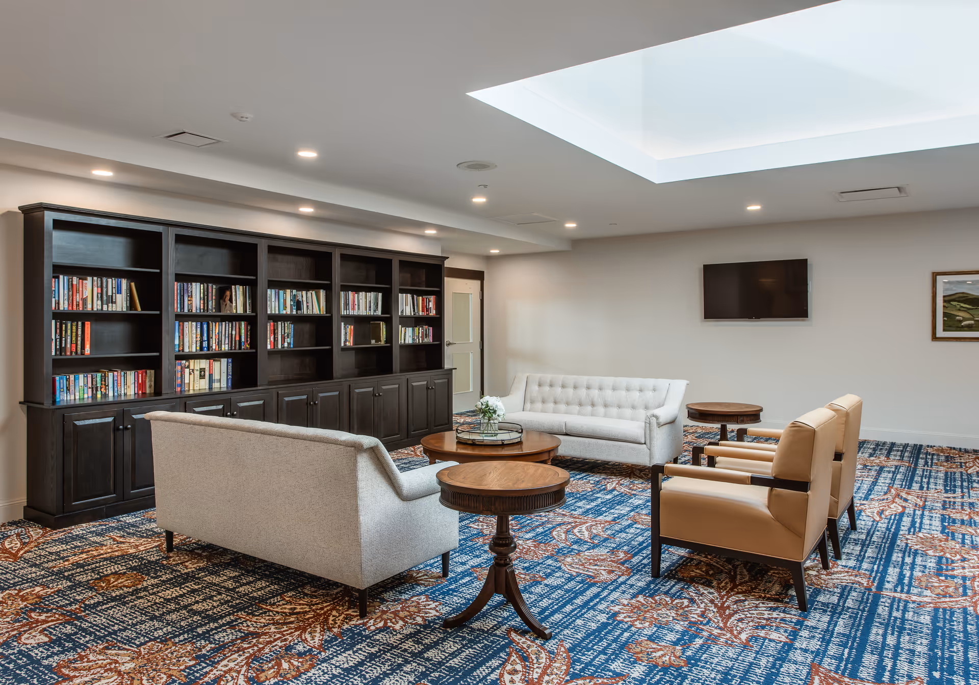 A spacious and well-lit common area with a large bookshelf filled with books along one wall. The room features two light gray sofas, two tan armchairs, and several round wooden tables arranged on a patterned blue and brown carpet. A flat-screen TV is mounted on the wall, and there is a skylight in the ceiling allowing natural light to brighten the space.