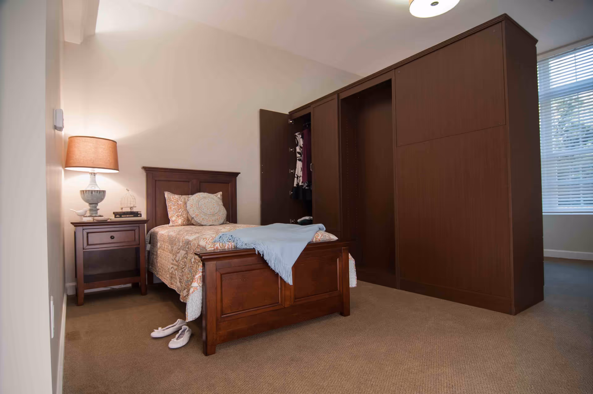 A cozy bedroom with a single wooden bed adorned with patterned bedding and decorative pillows. A light blue throw blanket is draped over the foot of the bed. Next to the bed is a wooden nightstand with a lamp, books, and decorative items. Behind the bed is a large wooden wardrobe with one door open, revealing hanging clothes. A window with blinds is visible on the right side, letting in natural light.