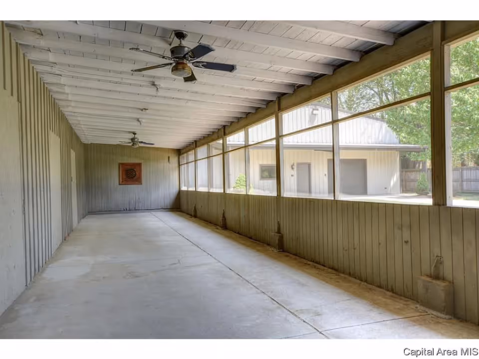 A long, enclosed porch or sunroom with a concrete floor, wooden paneled walls, and a white wooden ceiling with two ceiling fans. Large screened windows line one side, allowing a view of an adjacent building and trees outside.