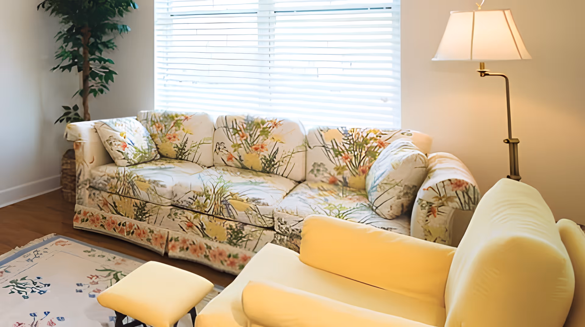 A cozy living room with a floral patterned sofa, a yellow armchair, a matching yellow ottoman, a floor lamp with a white shade, a potted plant in the corner, and a window with white blinds letting in natural light.
