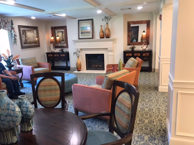 A cozy living room area in a senior living facility featuring several upholstered chairs in pink and orange hues arranged around a fireplace. The room has patterned carpet, framed artwork on the walls, decorative vases on the mantel and side tables, and a round wooden table with chairs in the foreground. Soft lighting and a window with floral curtains add to the warm ambiance.