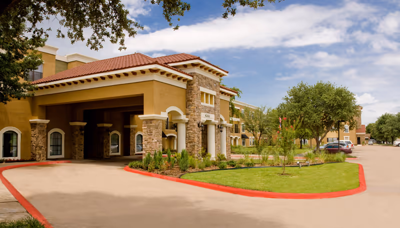 Front entrance of an assisted living facility with a porte-cochere, stone columns, driveway, and landscaped lawn.