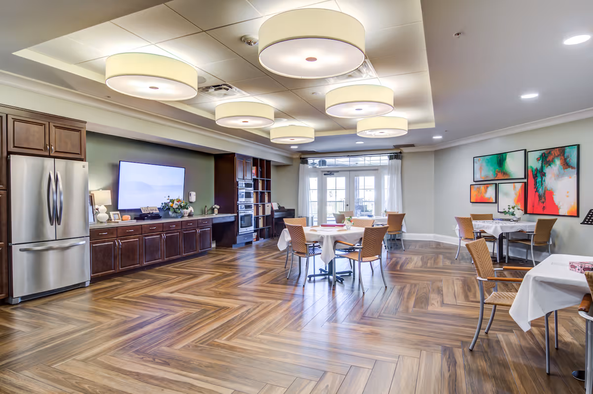 A spacious, well-lit dining area with wooden flooring and modern ceiling lights. The room features several tables with white tablecloths and wicker chairs. On the left side, there is a kitchen area with dark wooden cabinets, a stainless steel refrigerator, and built-in ovens. The far wall has large glass doors letting in natural light, and colorful abstract paintings hang on the right wall.