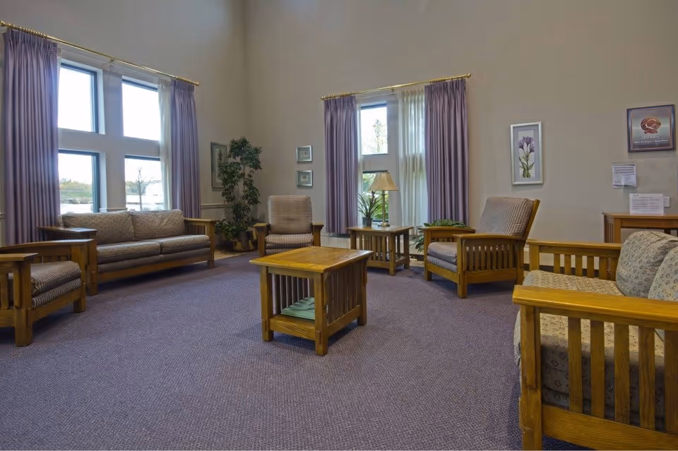 A spacious living room with wooden framed sofas and armchairs upholstered in patterned fabric, a wooden coffee table and side tables, purple curtains on large windows, a floor lamp, and framed artwork on the walls.