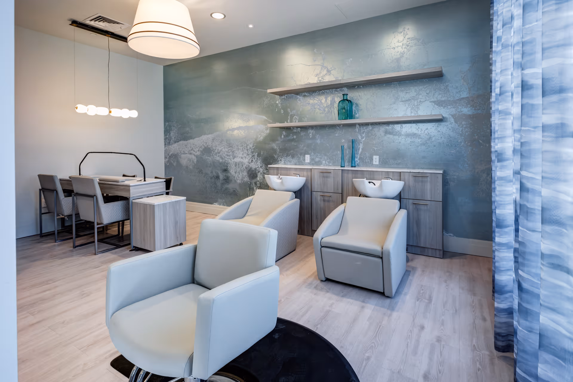 A modern salon area inside a senior living facility featuring two white salon chairs with wash basins, a light wood cabinet with shelves holding decorative glass vases, a dining table with four chairs, and contemporary lighting fixtures. The room has light wood flooring and a textured blue-gray accent wall.