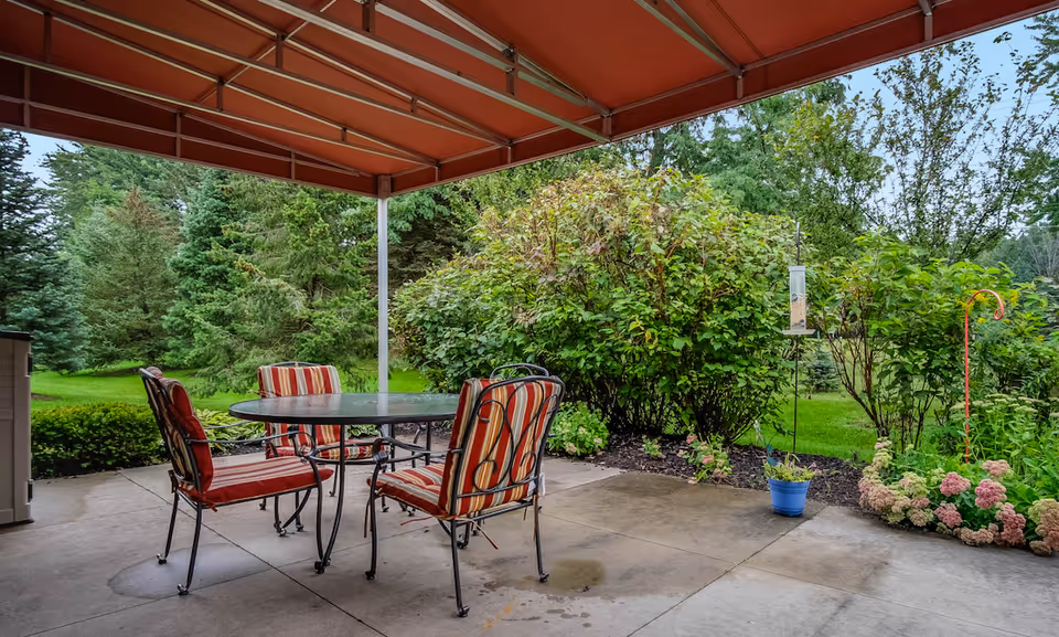 Outdoor patio area with a round glass table and four metal chairs with red and beige striped cushions under a red canopy. Surrounding the patio are green bushes, trees, and a well-maintained garden with flowers and a bird feeder.