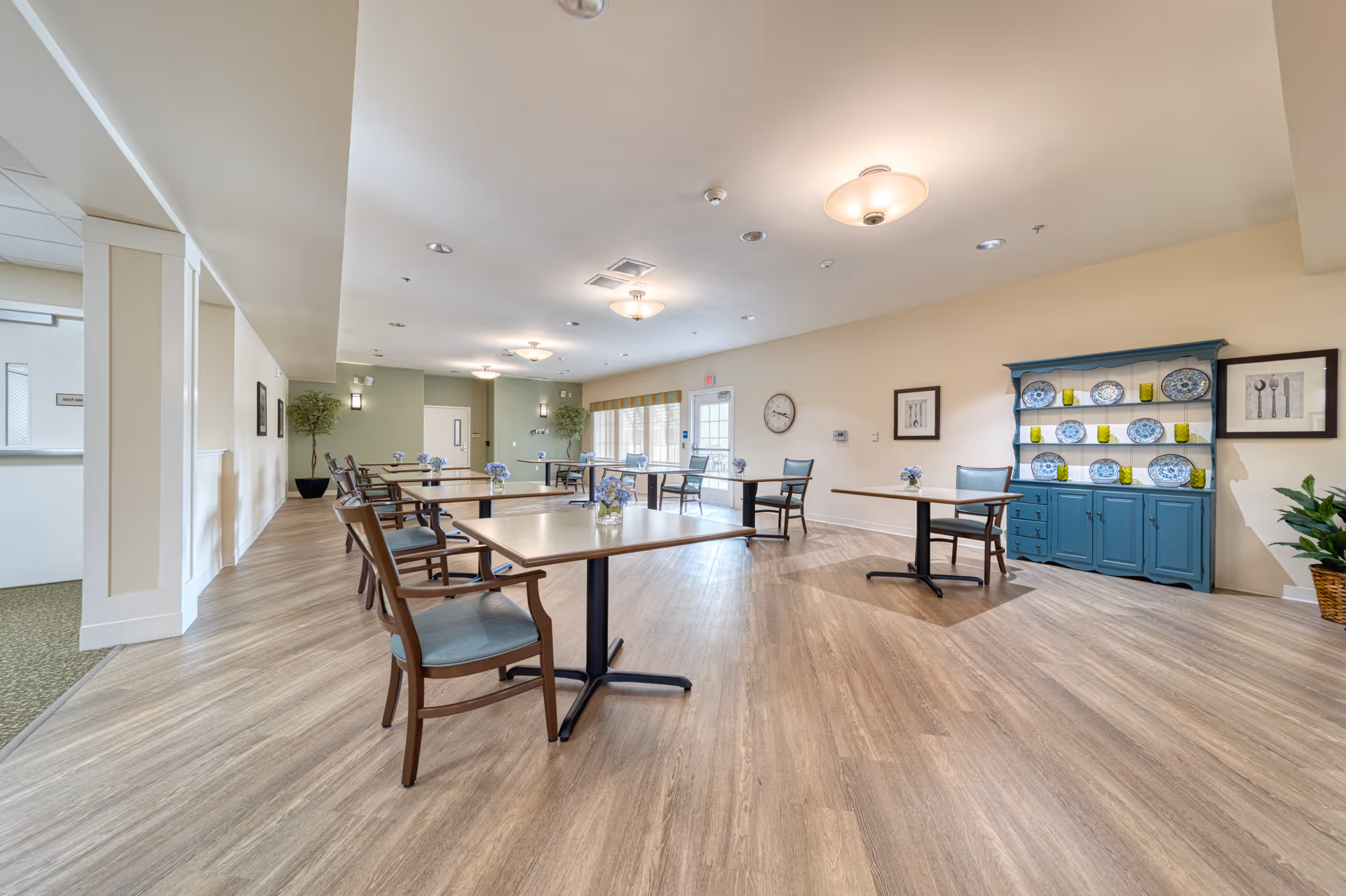 A spacious and well-lit dining room in a senior living facility with several tables and chairs arranged neatly. Each table has a small vase with purple flowers. The room features light wood flooring, cream-colored walls, a blue cabinet displaying decorative plates and glassware, framed artwork, and large windows letting in natural light.