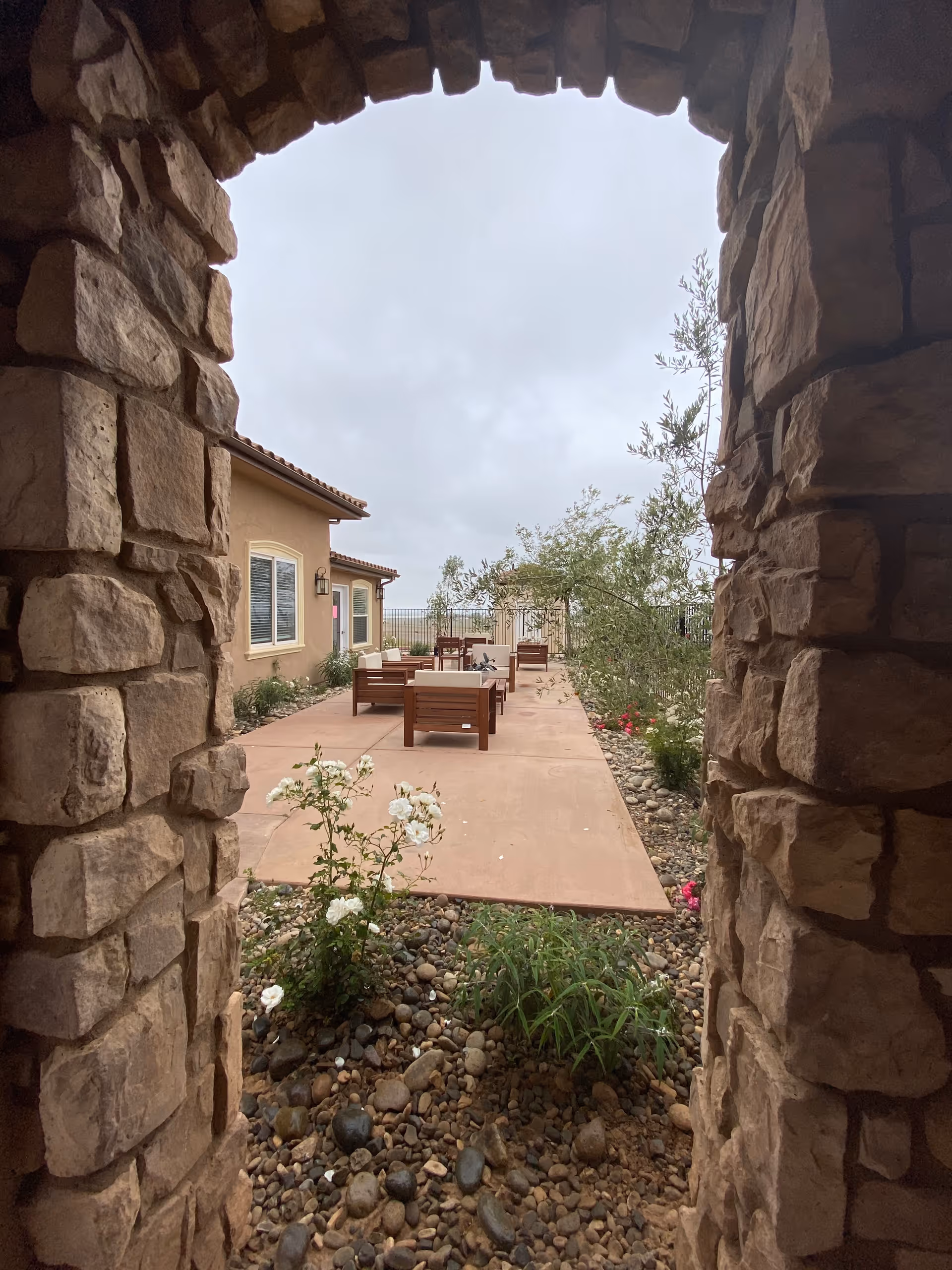 View through a stone archway looking out onto an outdoor patio area with wooden chairs and tables. The patio is surrounded by plants, small bushes, and a building with windows and a tiled roof. The sky is overcast.