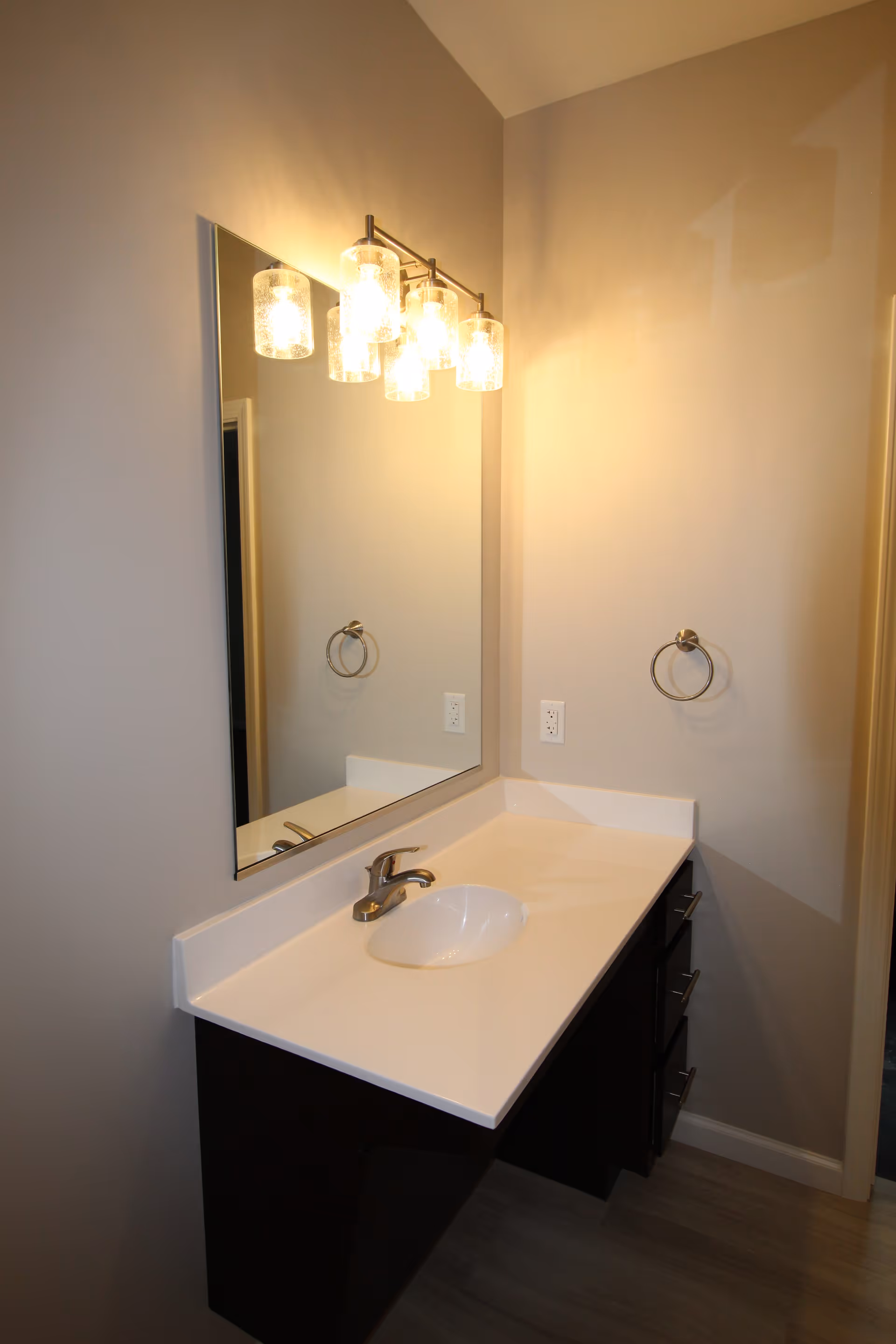 A bathroom vanity with a white countertop, an oval sink, and a silver faucet. Above the sink is a large rectangular mirror with a light fixture featuring five glass-covered bulbs. There is a towel ring mounted on the wall to the right of the mirror.