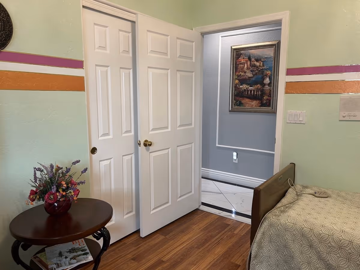 A small bedroom with light green walls featuring horizontal stripes in purple, white, and orange. There is a wooden floor, a bed with a patterned beige bedspread, and a small round wooden table with a vase of colorful flowers. Two white doors are partially open, revealing a hallway with gray walls, white trim, and a framed painting of a coastal scene.