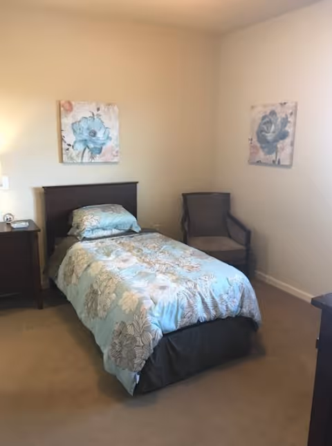 A small bedroom with a single bed covered in a blue floral comforter and matching pillow. There is a wooden nightstand with a lamp and a clock on the left side of the bed. A cushioned armchair is placed in the corner of the room. Two floral paintings hang on the beige walls above the bed and chair.