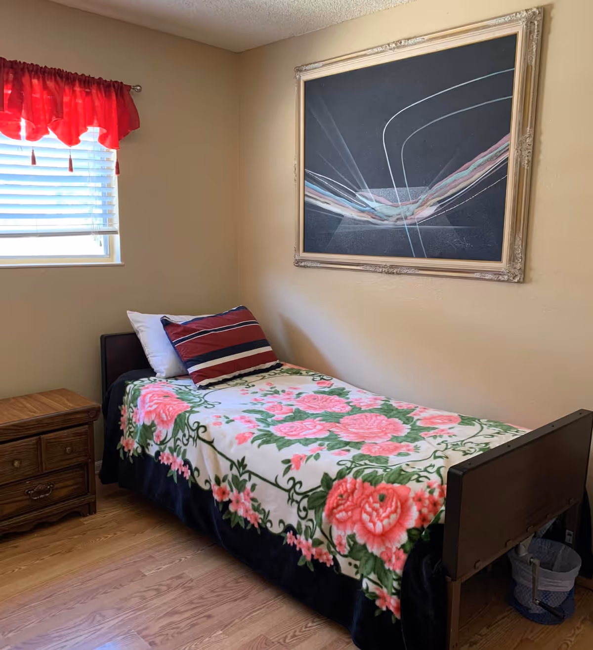 A small bedroom with a single bed covered with a floral blanket featuring pink roses and green leaves. There are two pillows on the bed, one white and one with red, white, and blue stripes. A wooden nightstand with drawers is next to the bed. A window with white blinds and a red valance is on the left wall. Above the bed, there is a framed abstract painting with curved lines on a dark background. The floor is wooden.