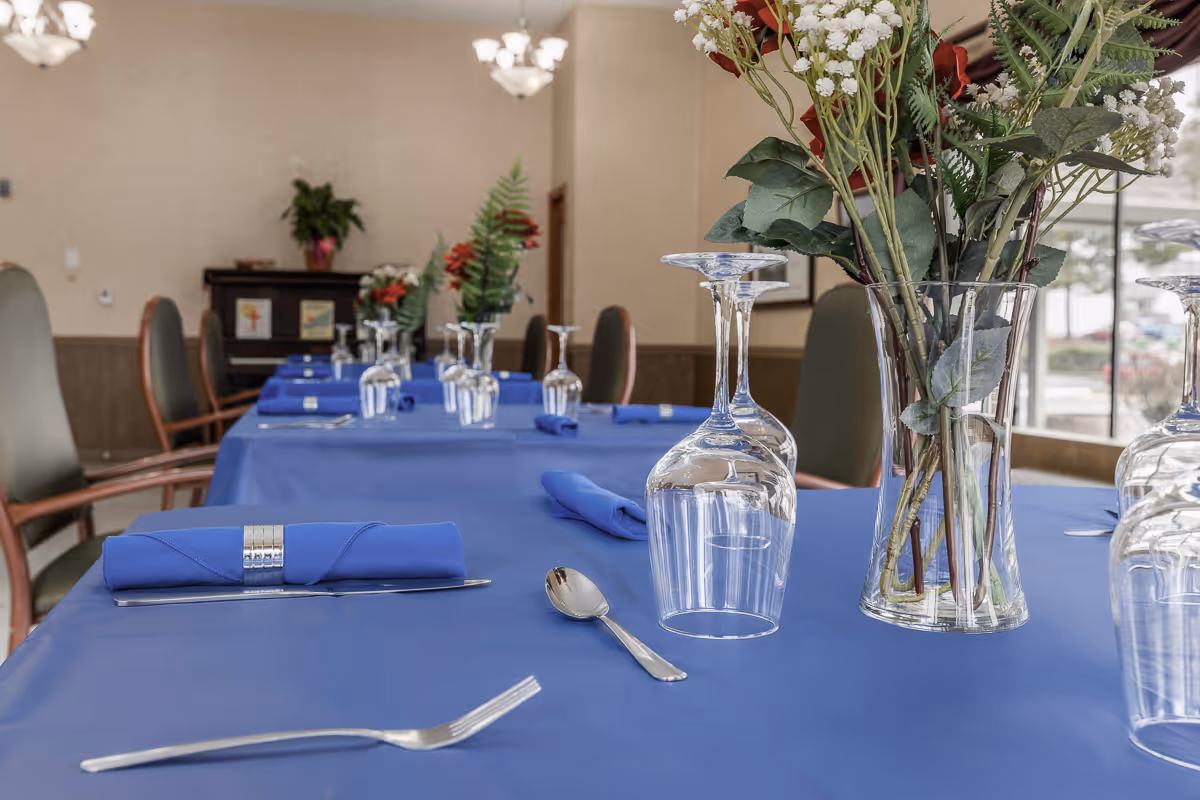 A dining table set with blue tablecloths, neatly folded blue napkins with silver napkin rings, upside-down wine glasses, and silverware including forks and spoons. A clear glass vase with red and white flowers is placed on the table. The background shows chairs and a piano against a beige wall with soft lighting fixtures.