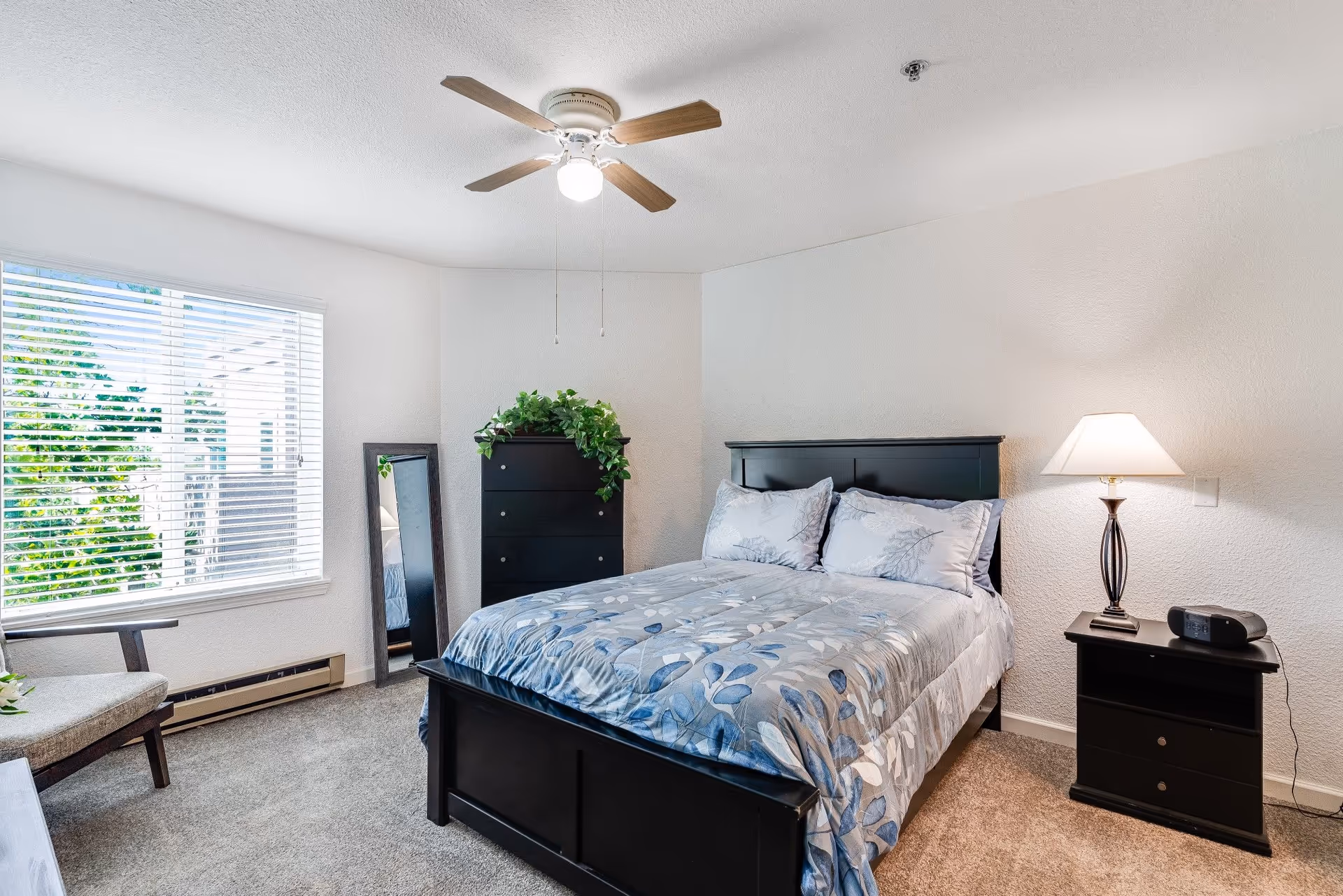 Sunlit bedroom featuring a bed with patterned bedding, nightstand and lamp, dresser with a plant, standing mirror, ceiling fan, and a large window with blinds.
