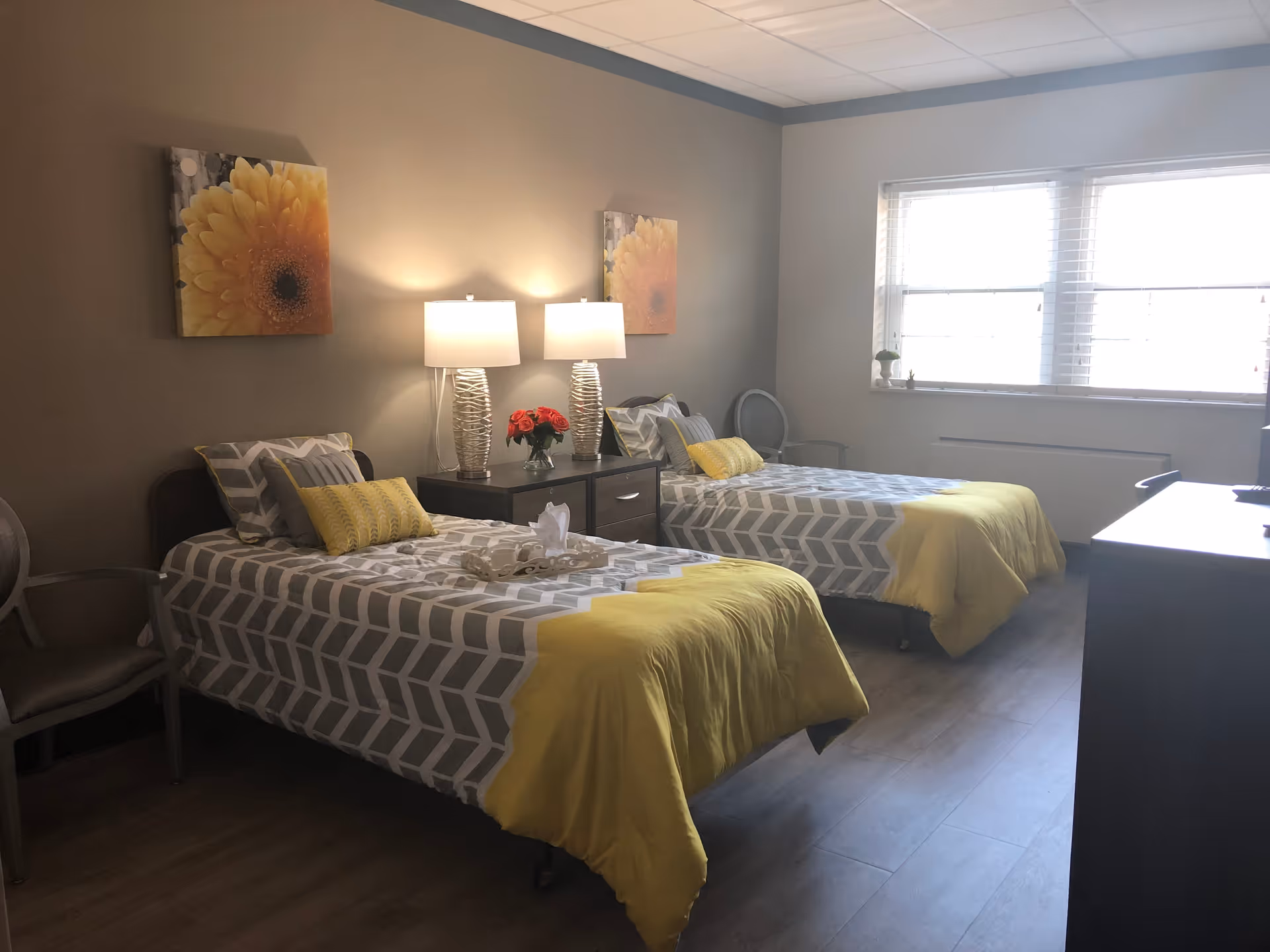 A bright and cozy bedroom with two single beds, each with gray and white patterned bedding and yellow blankets. Between the beds is a nightstand with two modern lamps and a vase of red flowers. The room has wooden flooring, a large window with blinds, two chairs, and wall art featuring large yellow flowers.