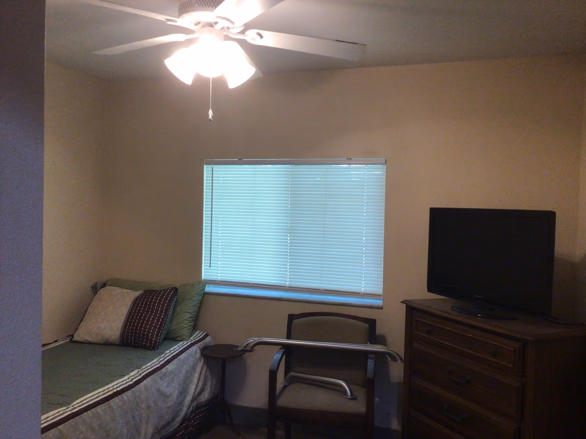 A small bedroom with a single bed against the left wall, a chair with metal support bars in front of a window with closed blinds, a wooden dresser with a flat-screen TV on top, and a ceiling fan with lights turned on.
