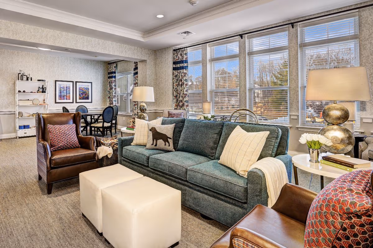 Sunlit communal living room with a teal sofa, leather armchairs, ottomans, lamps and a dining table by large windows.
