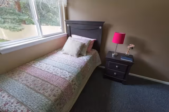A small bedroom with a single bed covered in a patterned quilt and two pillows, positioned next to a large window with a view of trees outside. Beside the bed is a dark wooden nightstand with two drawers, a pink lampshade, a small vase with pink flowers, and a book.