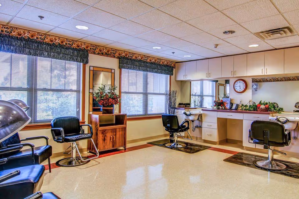 A bright and clean salon area inside a senior living facility with large windows covered by blinds and valances. The room features black salon chairs, hair dryers, a wooden cabinet with a mirror, and a counter with sinks and cabinets. Decorative plants and flowers are placed on the counter and cabinet.